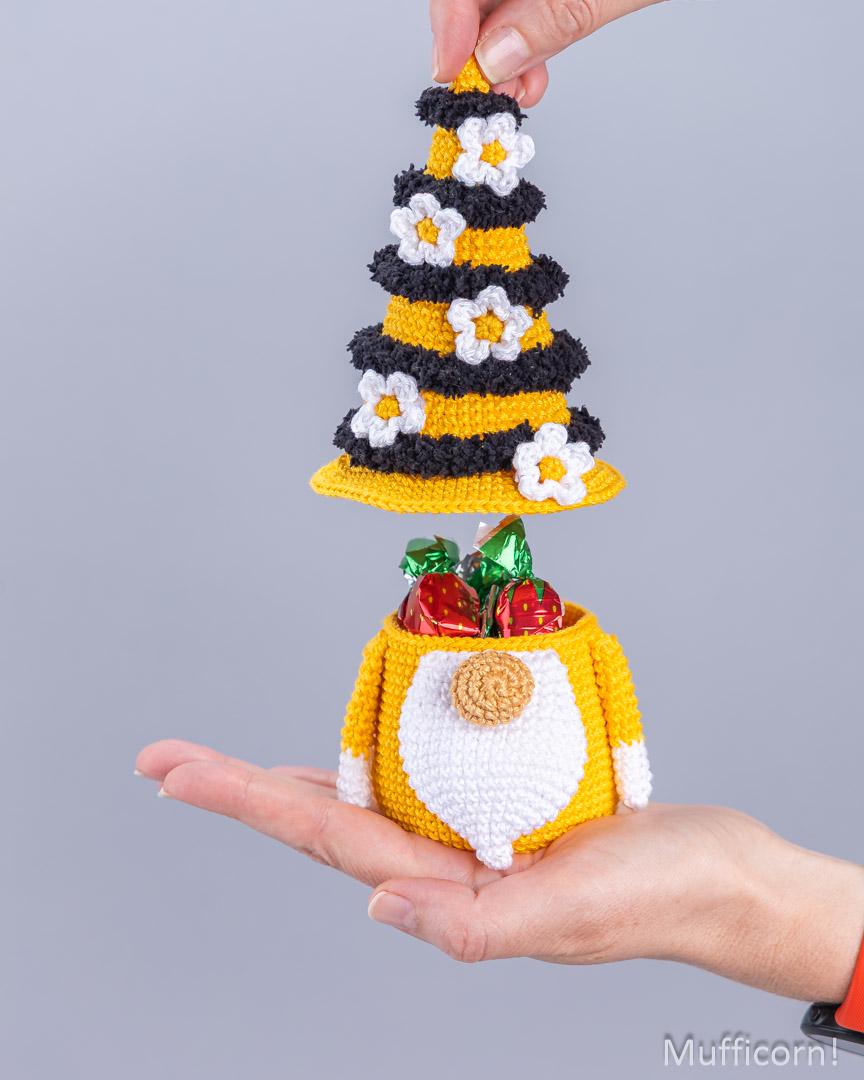 A small crocheted gnomehorn ornament with a yellow and black striped gnomehorn hat and a yellow-and-white gnomehorn body, shown being held in a person's hand against a plain gray background. The ornament is open to reveal wrapped chocolates inside, and the brand name Muffcorn is visible in the bottom right corner.