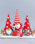 Three crocheted gnome decorations arranged on a white surface against a light gray background. The gnomes have red and white striped and solid red pointed hats decorated with white hearts, white beards, gray bodies, and red and white striped arm bands, with one holding a red heart.