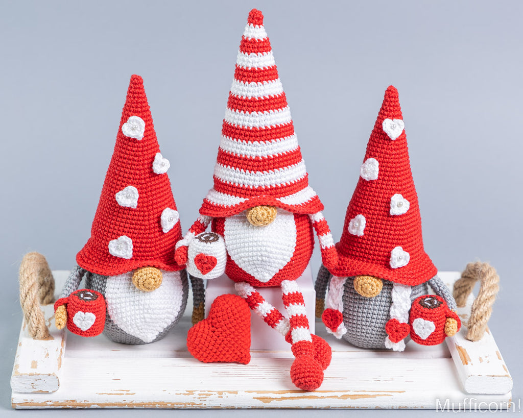 Three crocheted Valentine's Day gnomes arranged on a distressed white wooden tray against a light gray background. The gnomes have red and white striped hats, white beards, gray bodies, and red hearts and accessories.