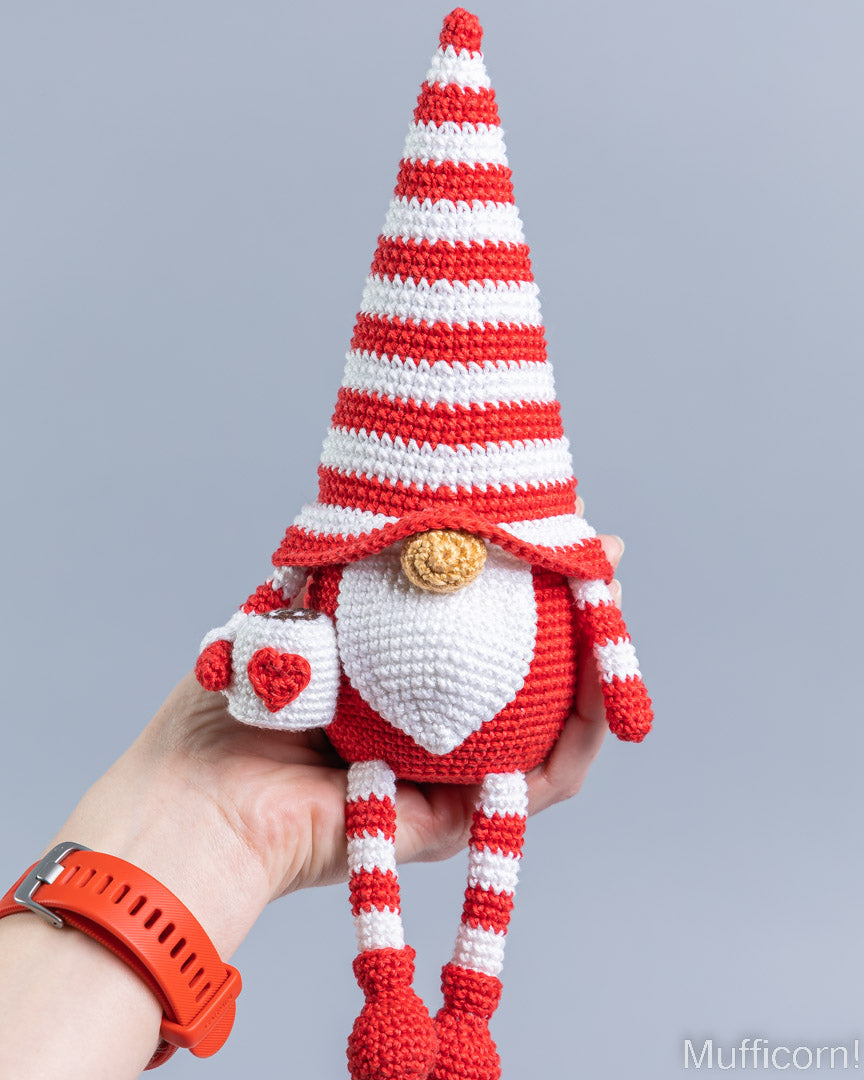 A crocheted gnome doll with a red and white striped hat, white beard, and red-and-white striped legs, being held in a person's hand against a plain light gray background.