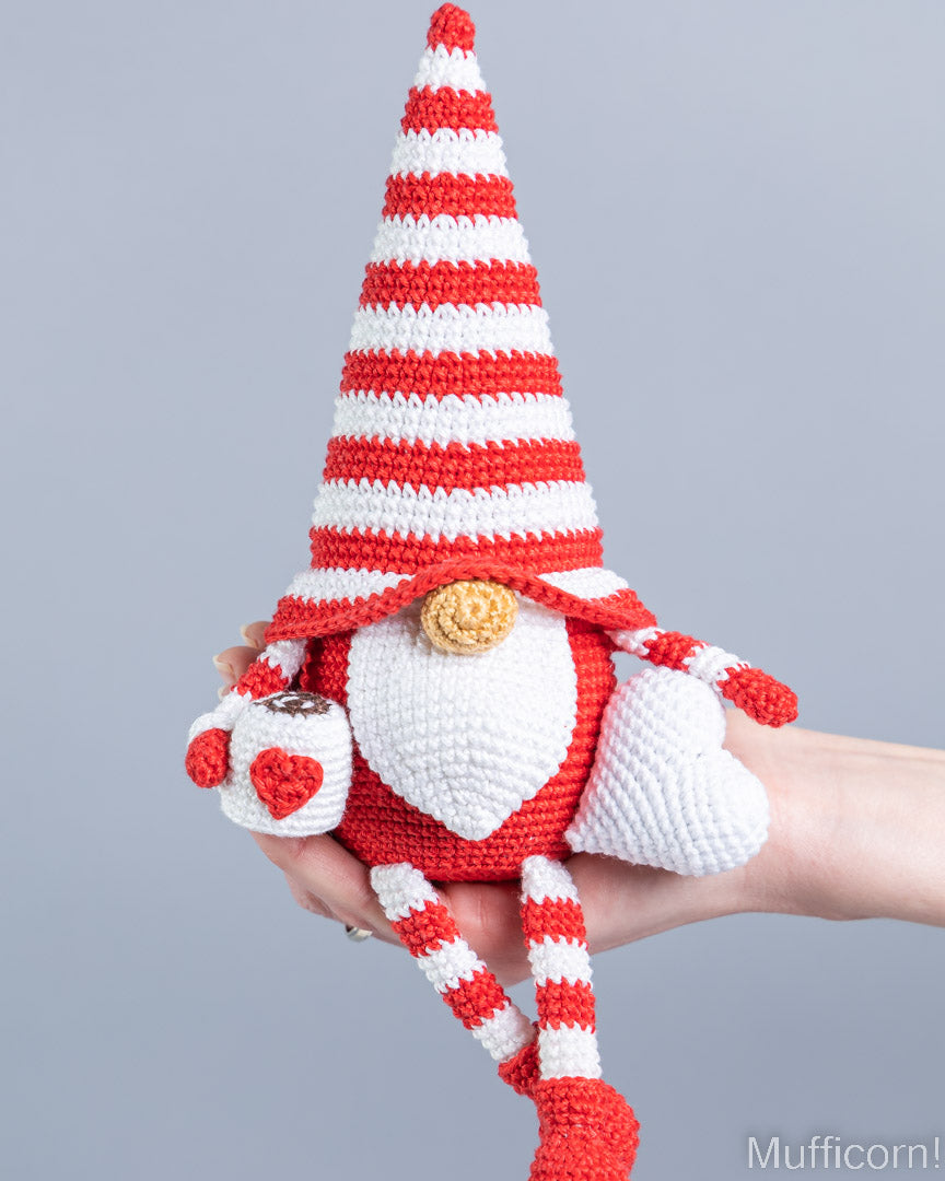 A crocheted gnome doll with cup and heart and a tall red-and-white striped hat, white beard, and red-and-white striped legs, being held in a person's hand against a plain light gray background.