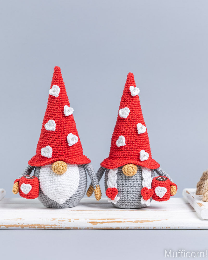 Two crocheted gnome decorations sitting side by side on a white surface against a light gray background. Each gnome has a red pointed hat with white heart details, a white beard, a gray body, and red-and-white heart-themed pockets.