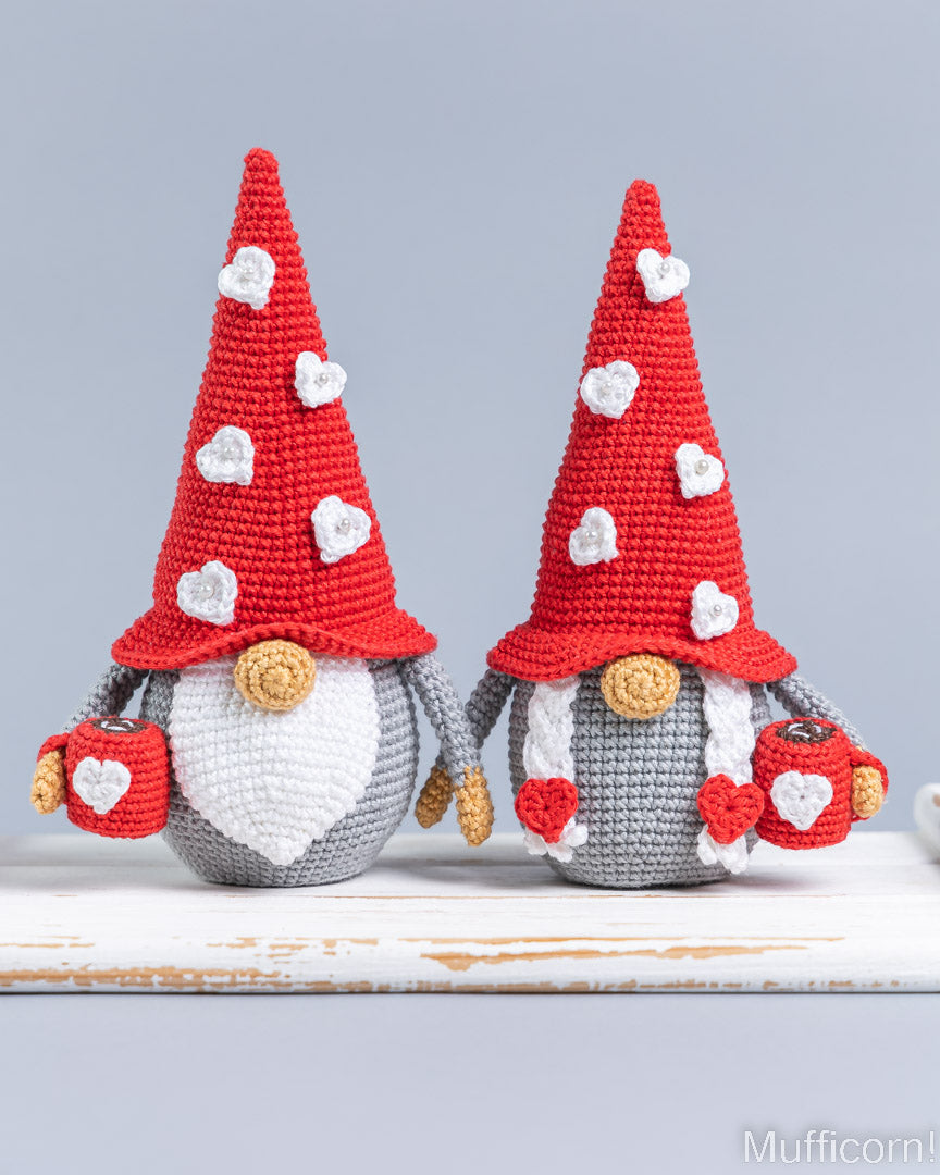 Two crocheted gnome figurines with red pointed hats decorated with white hearts, white beards, gray bodies, and small red-and-white heart accessories, displayed on a white surface against a light gray background.