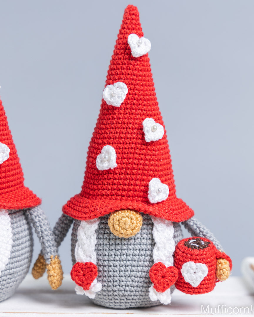 A crocheted gnome decoration with a tall red hat decorated with white hearts, a gray body, white beard, and a small red mug with a heart, shown next to another similar gnome against a light gray background.