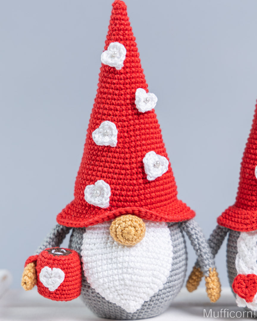 A crocheted gnome doll with a tall red hat decorated with white hearts, a white beard, gray body, and a small red mug, shown against a soft light gray background with part of another gnome visible on the right.