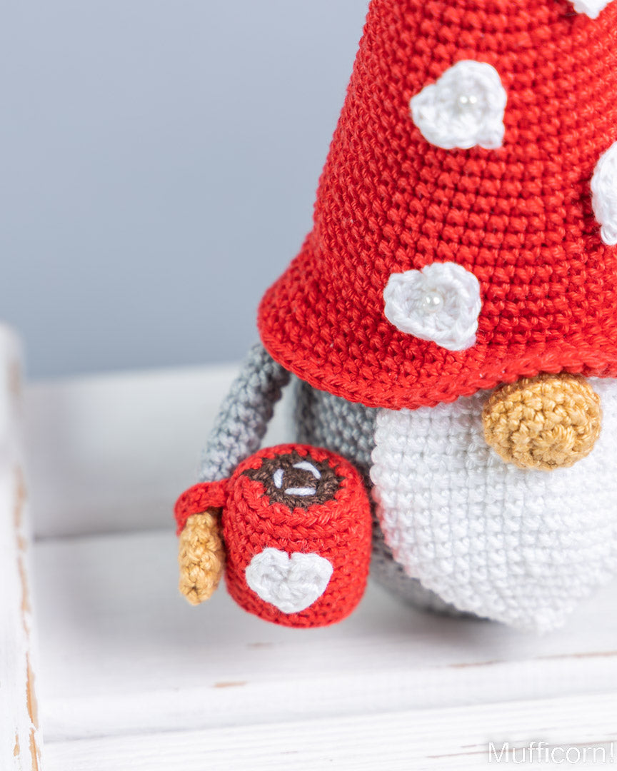 Close-up of a crocheted gnome doll with a red mushroom-shaped hat, white polka dots, gray arms, a white beard, and a small red mug with a white heart, set against a soft gray and white background.
