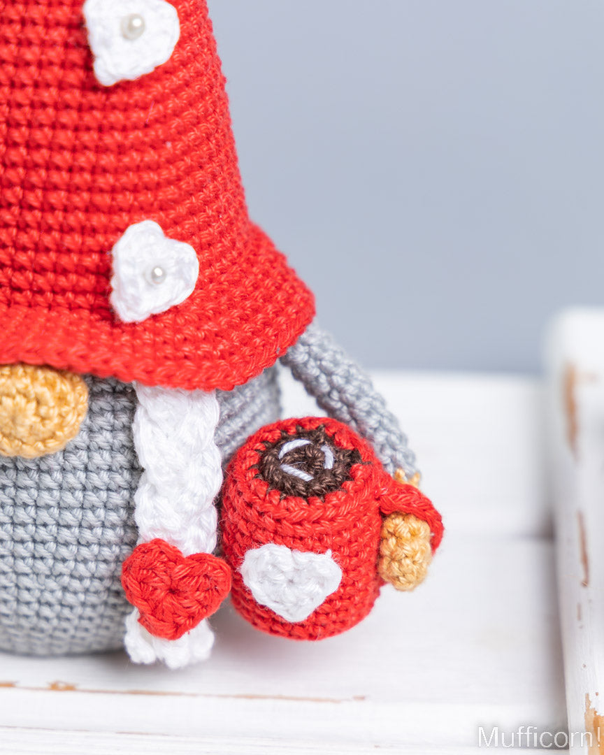 Close-up of a crocheted gnome doll wearing a red hat with white heart details, holding a red mug with a heart and a small red heart, on a white surface with a soft gray background.