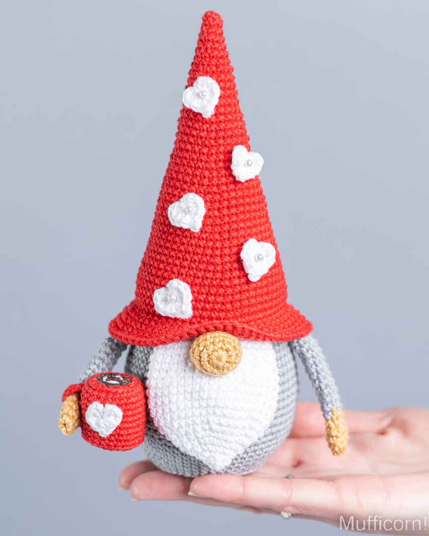 A crocheted gnome doll with a tall red hat decorated with white hearts, a white beard, gray body, and gold details, being held in a person's hand against a light gray background.