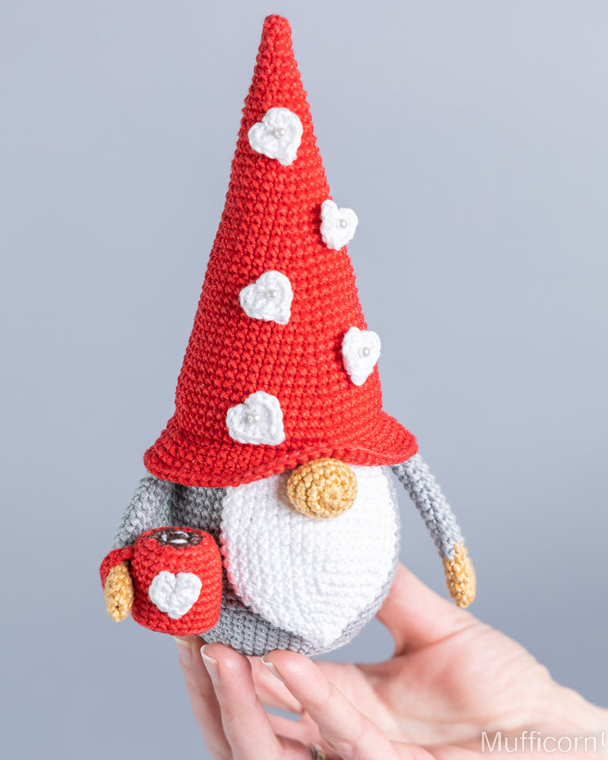 A crocheted gnome doll with a tall red hat decorated with white hearts, a white beard, gray body, and gold details, being held in two hands against a plain gray background.