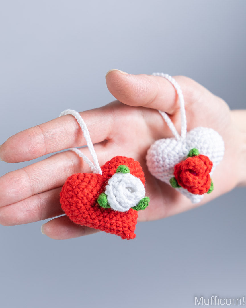 A hand holding two small crocheted heart-shaped ornaments, one red with a white rose and green leaves, and one white with a red rose and green leaves, against a soft gray background.