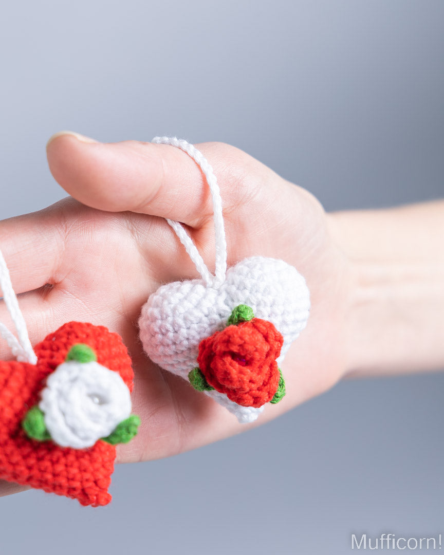 A hand holds two small crocheted heart-shaped ornaments, one white with a red rose and one red with a white rose, against a soft gray background.