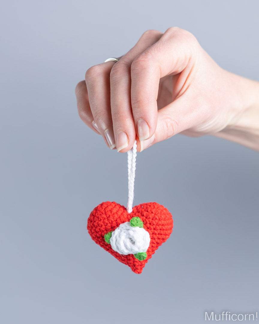 A hand holding a small crocheted red heart ornament with a white rose and green leaves, suspended by a white string, against a solid gray background.