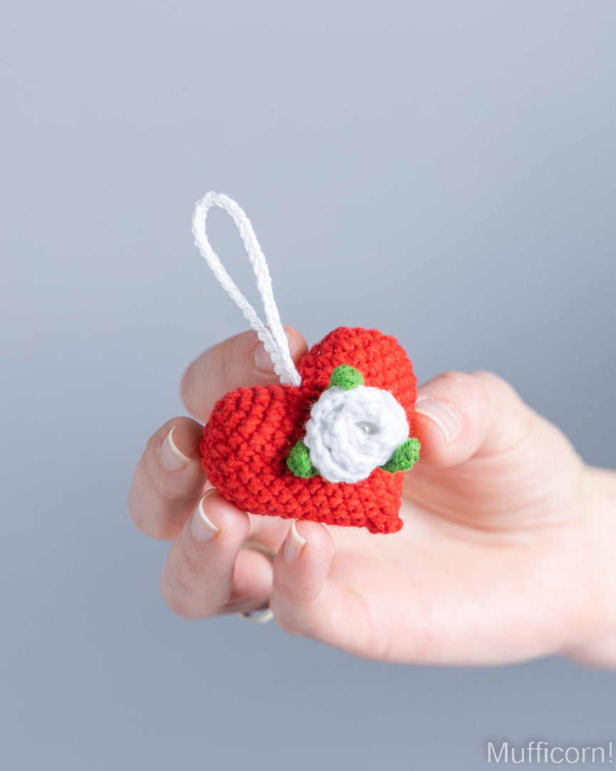 A hand holding a small crocheted red heart ornament with a white hanging loop and a white flower with green leaves, against a soft gray background.