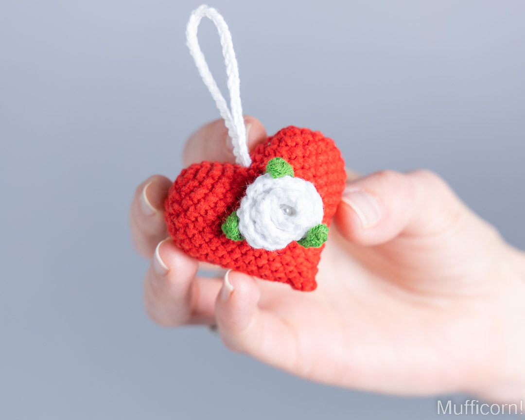 A hand holding a small crocheted red heart ornament with a white hanging loop, a white flower center, and green leaf accents against a soft gray background.