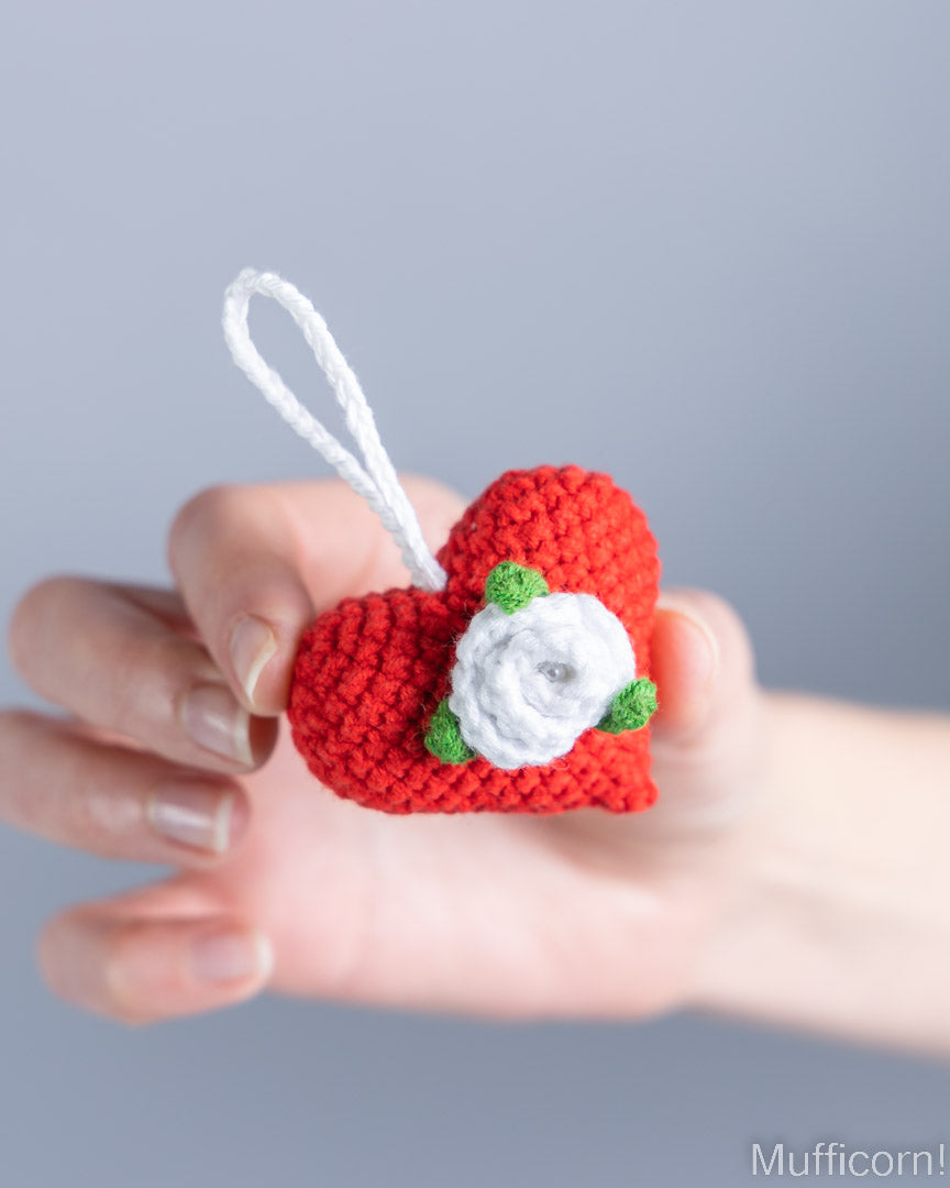 A hand holding a small crocheted red heart ornament with a white hanging loop, a white flower, and green leaves, against a soft gray background.