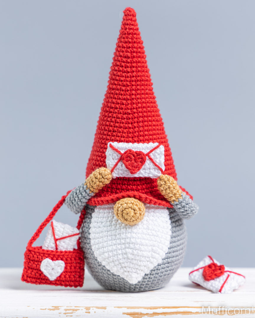 A crocheted gnome figurine with a tall red hat, white beard, gray body, and red-and-white envelope-themed accessories, shown on a light gray background with a small matching envelope at its side.