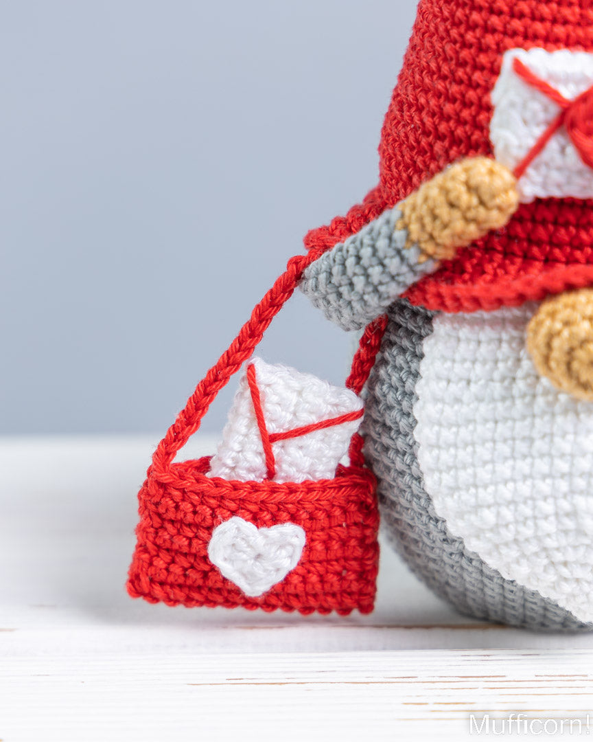 A close-up of a crocheted penguin figure wearing a red hat and carrying a small red pouch with a white heart and white envelope detail, set against a soft light gray background.
