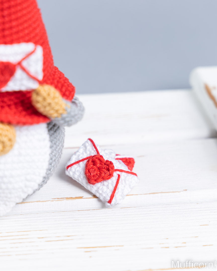 A close-up of a crocheted white gift box with a red heart-shaped bow and red ribbon, placed on a white wooden surface next to a partially visible red and white crocheted figure.