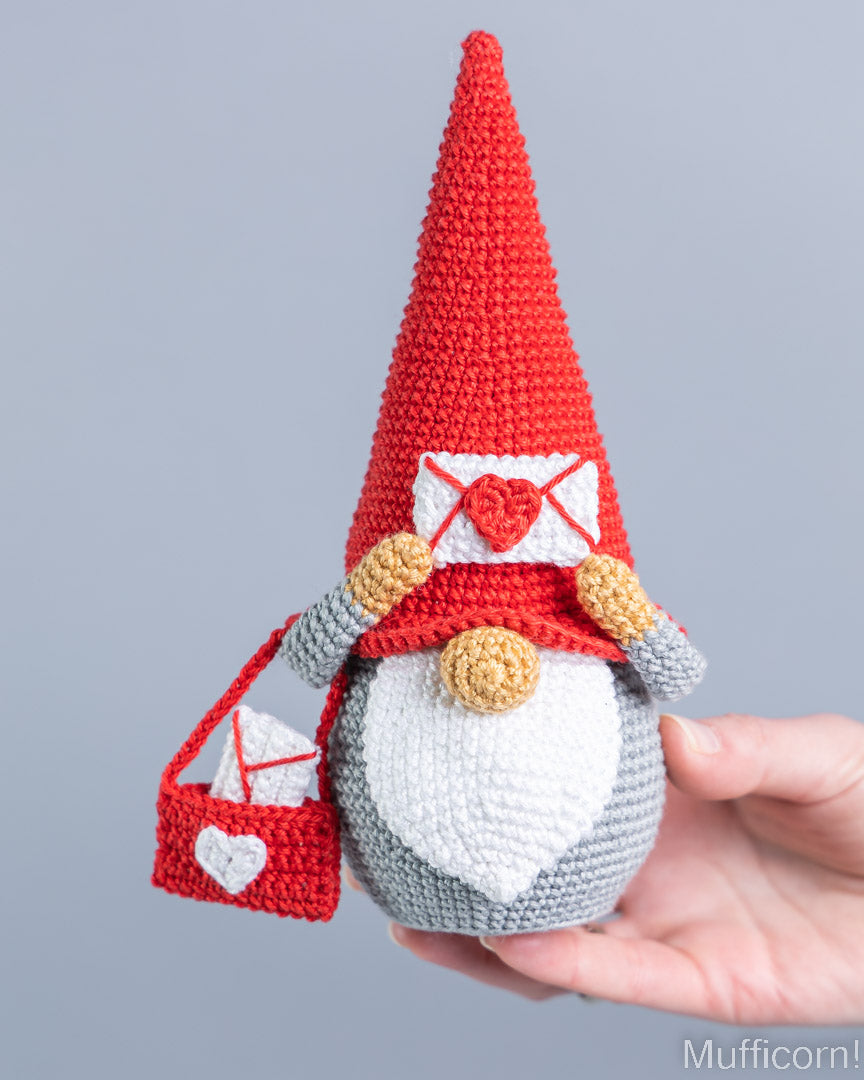 A crocheted gnome doll with a tall red hat, white beard, and red-and-white heart-themed accessories, being held in a person's hand against a plain light gray background.