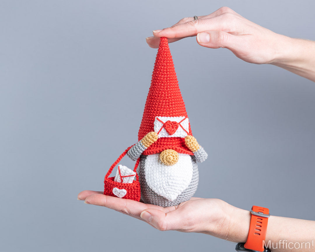 A crocheted gnome ornament being held in two hands against a plain gray background. The gnome has a tall red hat, a white beard, gray body, and carries a red sack with white hearts.