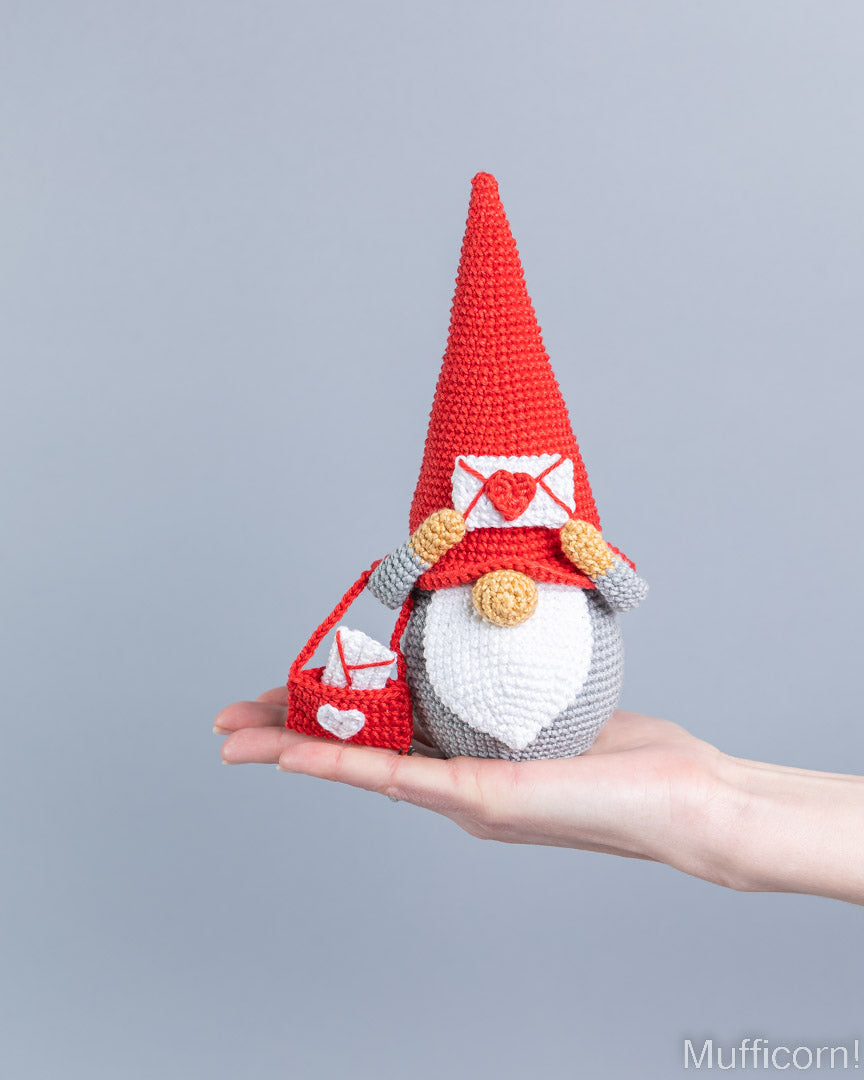 A crocheted gnome doll with a tall red hat, white beard, and red-and-white mailbox, being held in a person's hand against a plain light gray background.