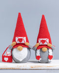 Two crocheted gnomes with tall red hats, white beards, gray bodies, and red-and-white heart details, displayed on a rustic white surface against a light gray background.