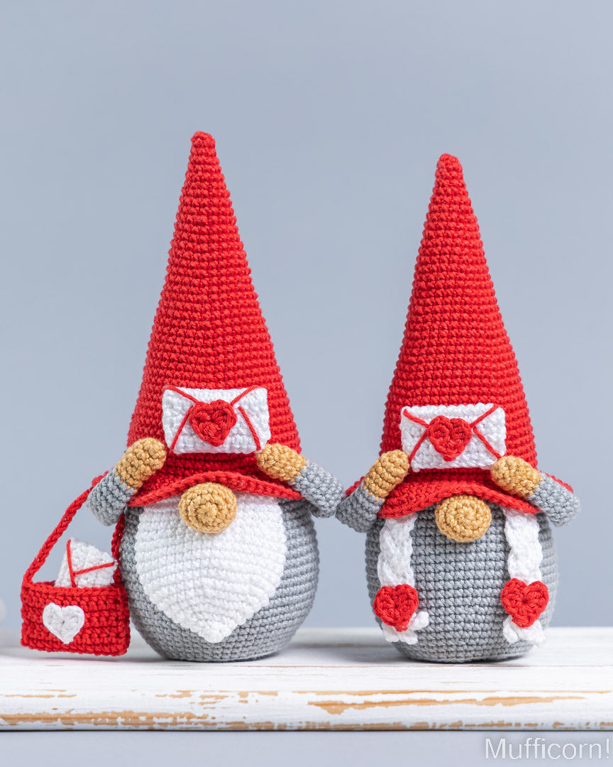 Two crocheted gnomes with tall red hats, white beards, gray bodies, and red-and-white heart details, displayed on a rustic white surface against a light gray background.