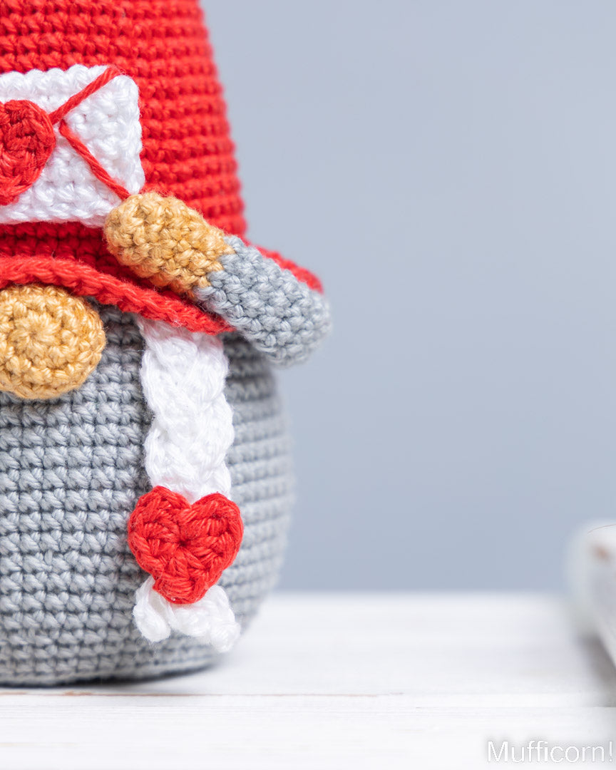 Close-up of a crocheted gnome figurine wearing a red hat with a white envelope and red heart detail, a gray sleeve, and a red heart hanging from its side, set against a soft gray background.