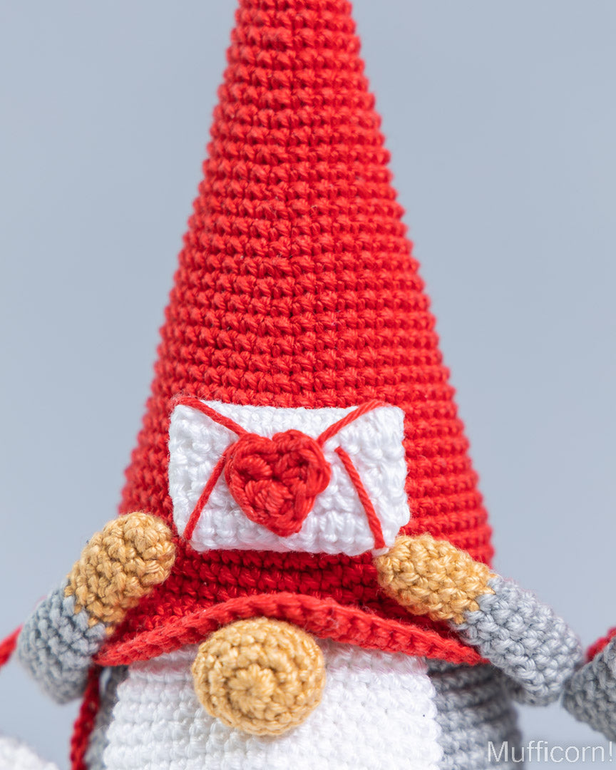 Close-up of a crocheted gnome doll wearing a tall red pointed hat with a white envelope and red heart detail on the front, plus gray and white body sections and tan accents, against a light gray background.