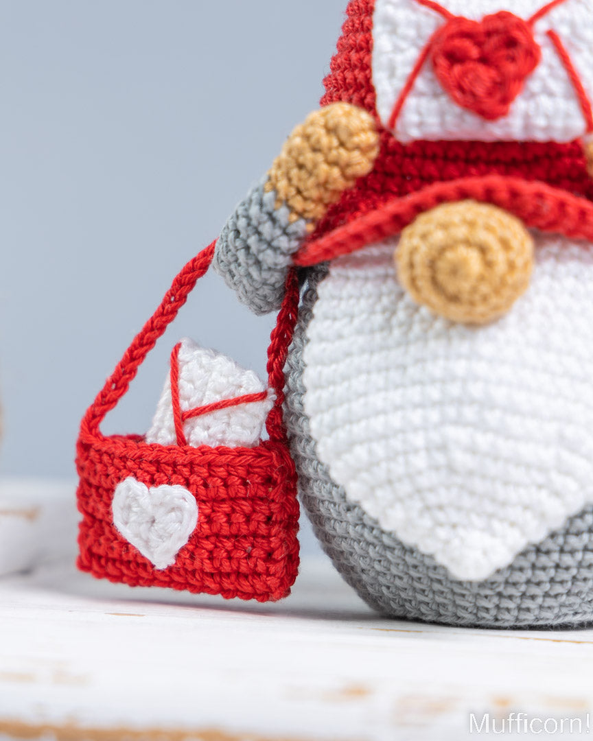 A close-up of a crocheted gnome figure in red, white, and gray, holding a small matching red and white crocheted gift bag with a white heart detail, against a soft light blue background.