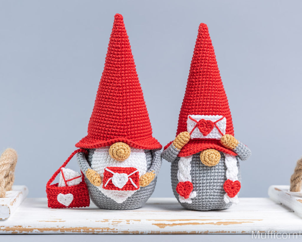 Two crocheted gnomes with tall red hats, gray bodies, and red-and-white envelopes, displayed on a white rustic surface against a light gray background.
