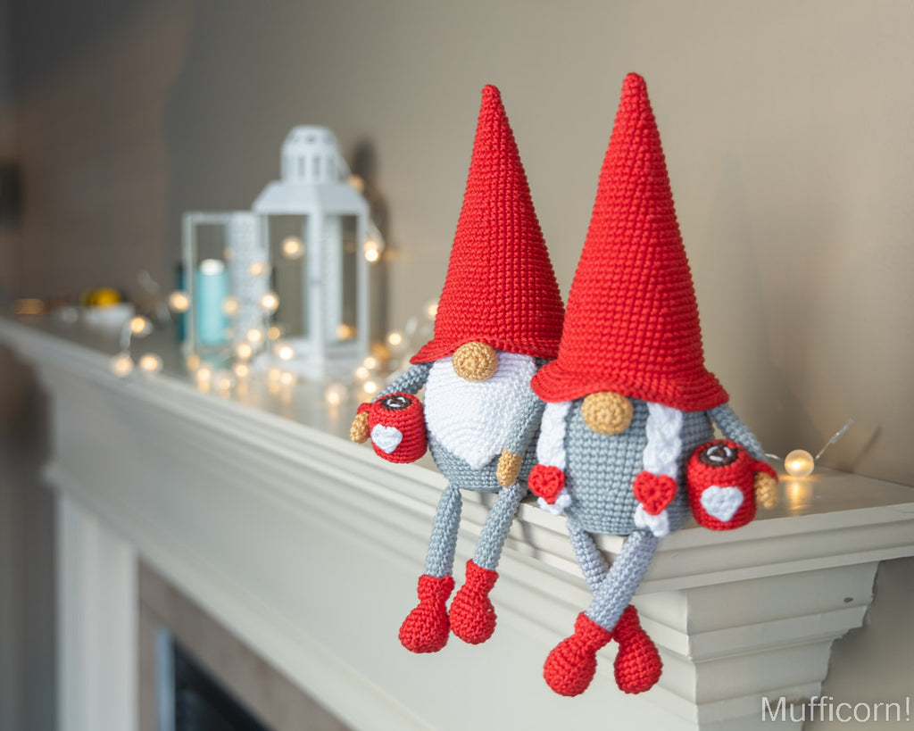Two crocheted gnome decorations with tall red hats, gray bodies, and red shoes hanging from a white mantel decorated with warm fairy lights and a white lantern in the background.