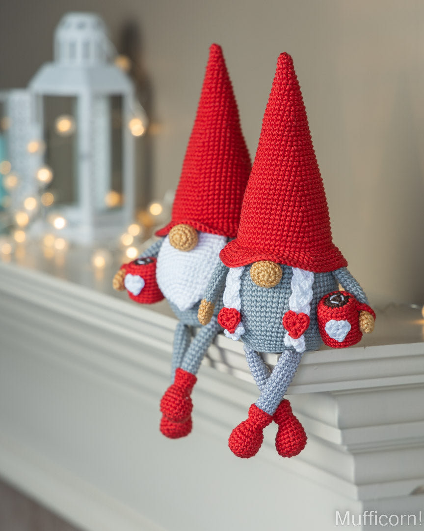 Two crocheted garden gnome dolls with tall red hats, white beards, gray bodies, and red shoes, posed on a white mantel with a blurred lantern and warm string lights in the background.