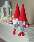 Two crocheted garden gnome dolls with tall red hats, white beards, gray bodies, and red shoes, posed on a white mantel with a blurred lantern and warm string lights in the background.