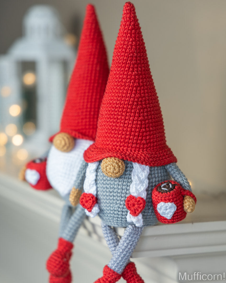 A close-up of a crocheted gnome decoration with a tall red hat, white beard, gray body, and red-and-white heart details, shown leaning on a white surface with a softly blurred indoor background.