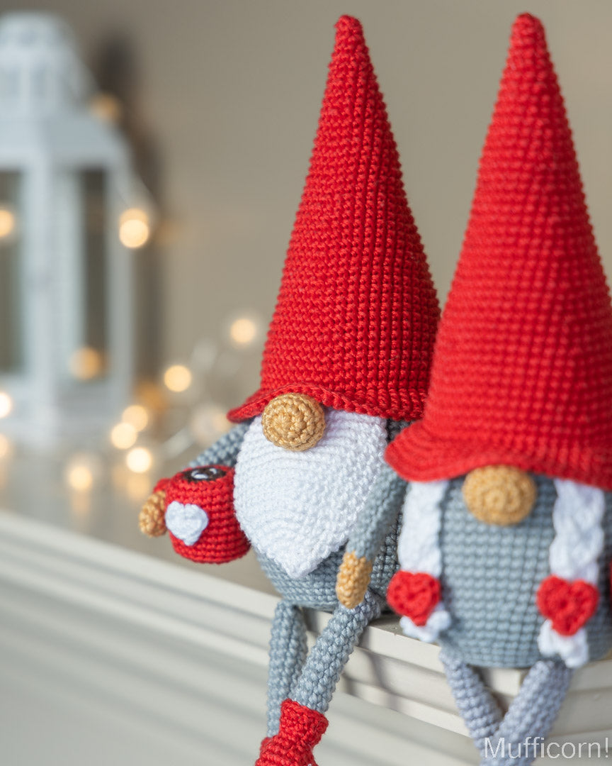 Two crocheted Christmas gnomes with tall red hats and gray bodies, posed on a windowsill with warm blurred lights in the background.