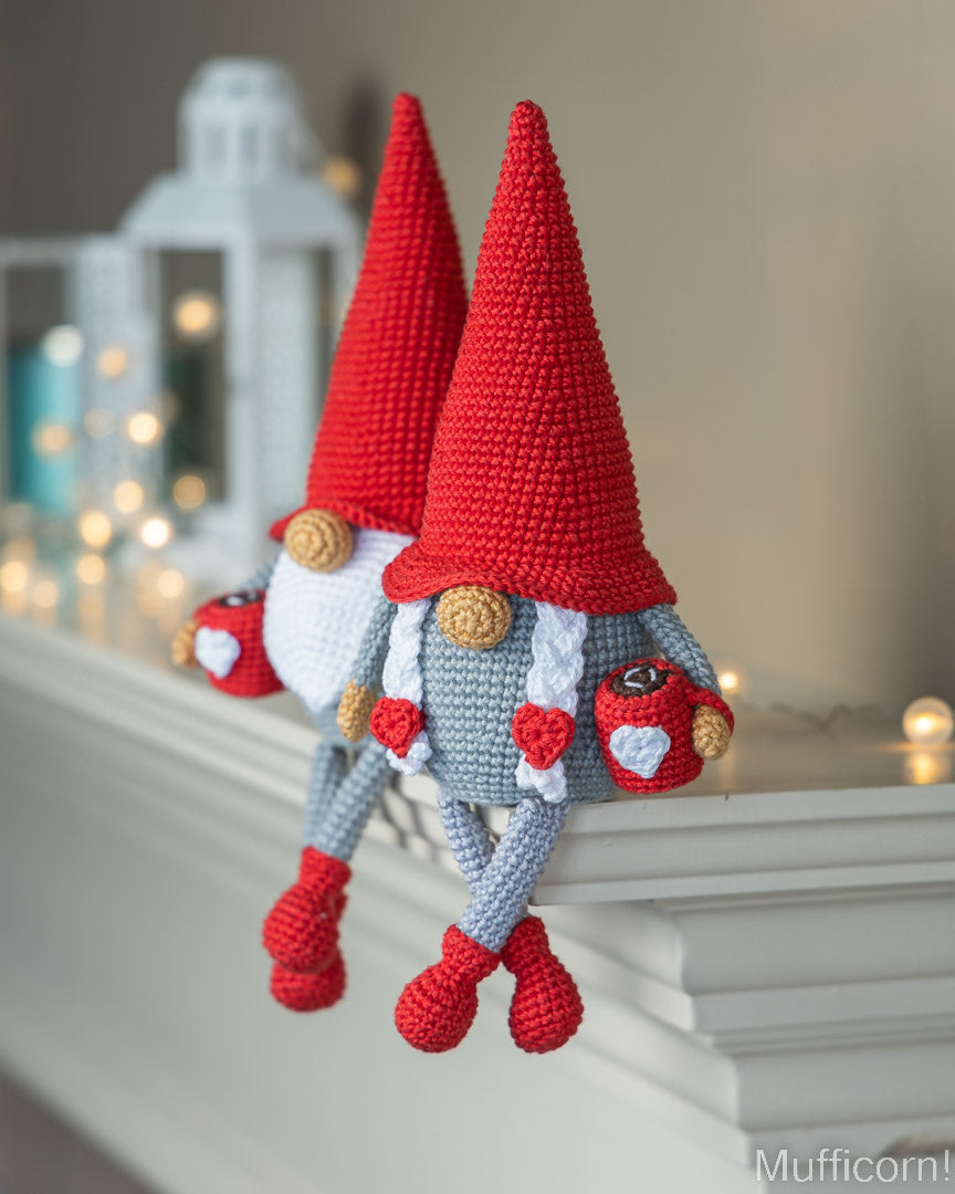 A crocheted gnome doll with a tall red hat, white beard, gray body, and red shoes, posed on a white mantelpiece in a softly lit indoor setting.