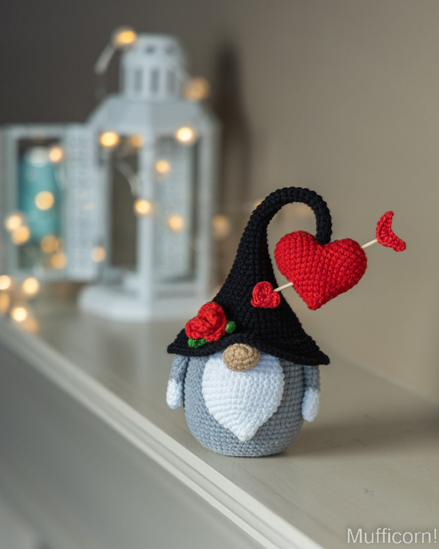 A crocheted gnome figurine with a black hat, red heart-shaped arrow, red roses, and a white beard, displayed on a shelf in front of a softly blurred white lantern with warm lights.