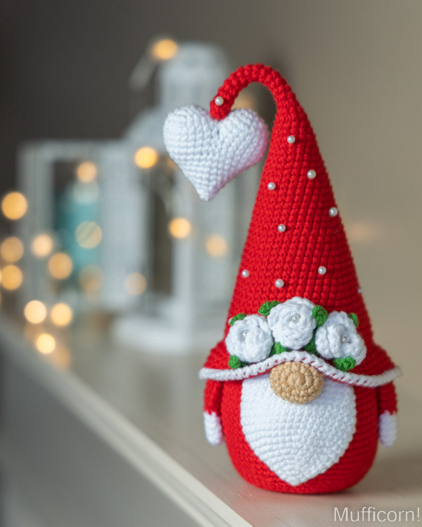 A crocheted Christmas gnome decoration with a tall red hat, white heart-shaped face, white roses, green leaves, and a small beige nose, displayed on a shelf with blurred warm lights in the background.