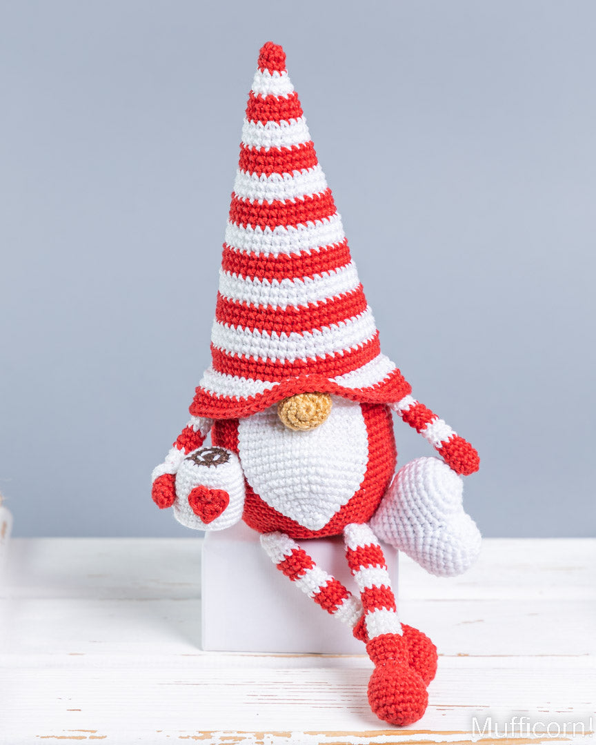 A crocheted gnome doll with heart and cup a tall red-and-white striped hat, white belly, and red-and-white striped legs, posed sitting on a white block against a light gray background.