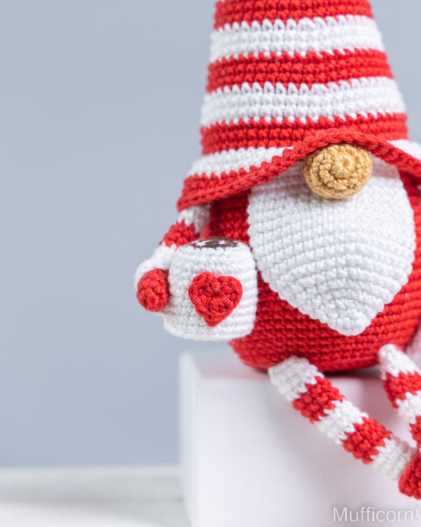 A close-up of a crocheted cup of a gnome with red and white striped hat and legs, a white beard, and a small tan button, holding a white mug with red hearts. The background is a soft light gray with a white pedestal.