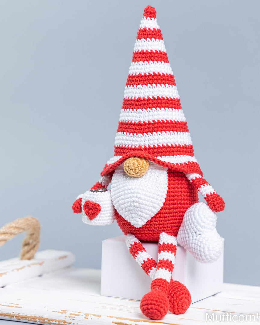 A crocheted gnome doll with a tall red-and-white striped hat, white beard, red body, and striped legs, posed sitting on a white block against a light gray background.