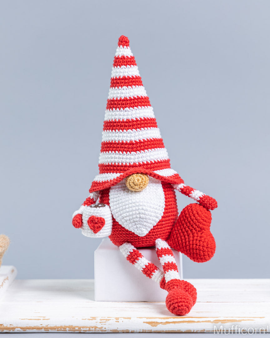 A crocheted gnome doll with a tall red-and-white striped hat, white beard, red nose, and red-and-white striped legs, posed on a white block against a light gray background.