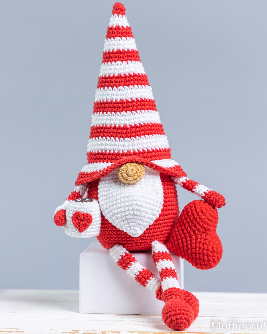 A crocheted gnome doll with a red and white striped hat, white beard, and red body, sitting on a small white block against a light gray background.