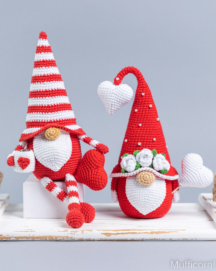 Two crocheted Valentine gnomes in red and white, displayed on a white wooden surface against a light gray background.