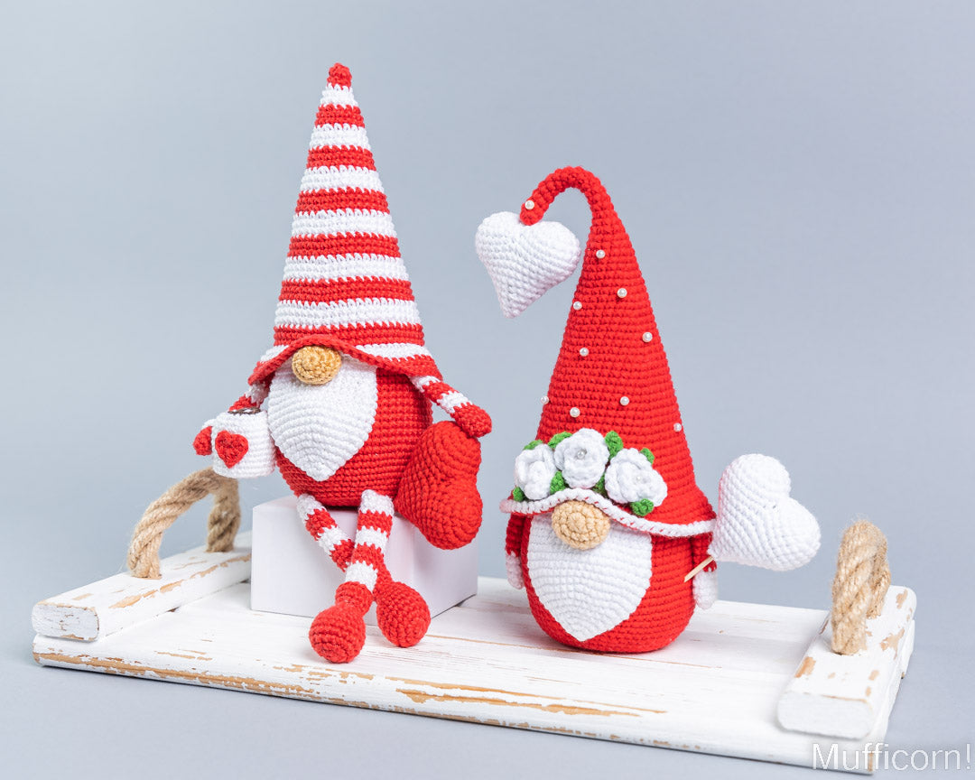 Two crocheted Valentine gnomes in red and white, displayed on a distressed white wooden tray against a light gray background.