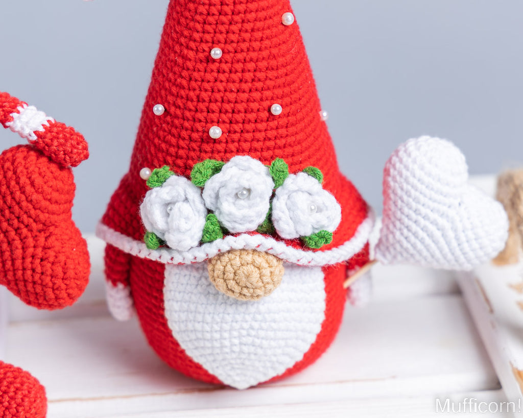 A crocheted Valentine gnome doll with a red pointed hat decorated with small white pearl-like beads, a white beard, white roses with green leaves on the forehead, and a white heart-shaped hand, shown on a light tabletop.