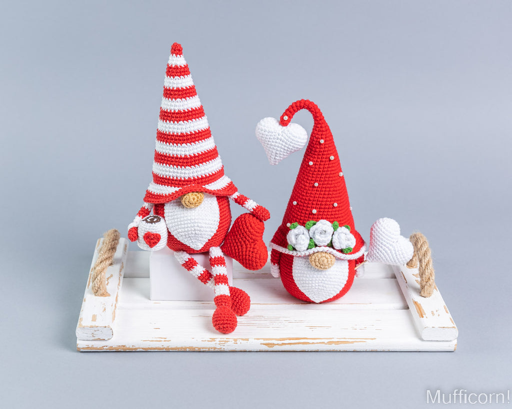 Two crocheted Valentine gnomes sitting on a distressed white wooden tray against a light gray studio background. One gnome wears a red-and-white striped hat and red-and-white striped pants, while the other wears a solid red hat with white accents.