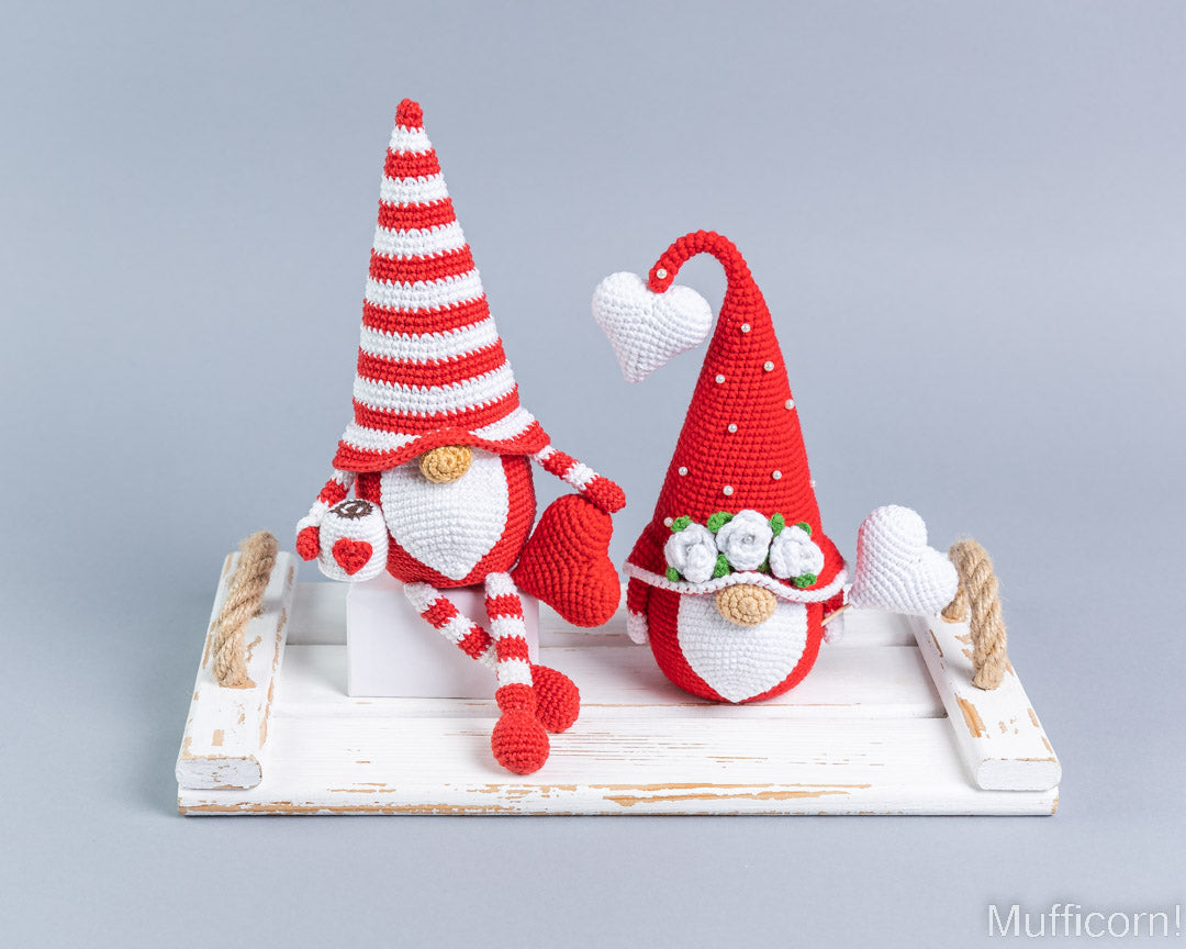 Two crocheted Valentine gnomes sitting on a distressed white wooden tray against a light gray studio background. One gnome wears a red-and-white striped hat and red-and-white striped pants, while the other wears a solid red hat with white accents.