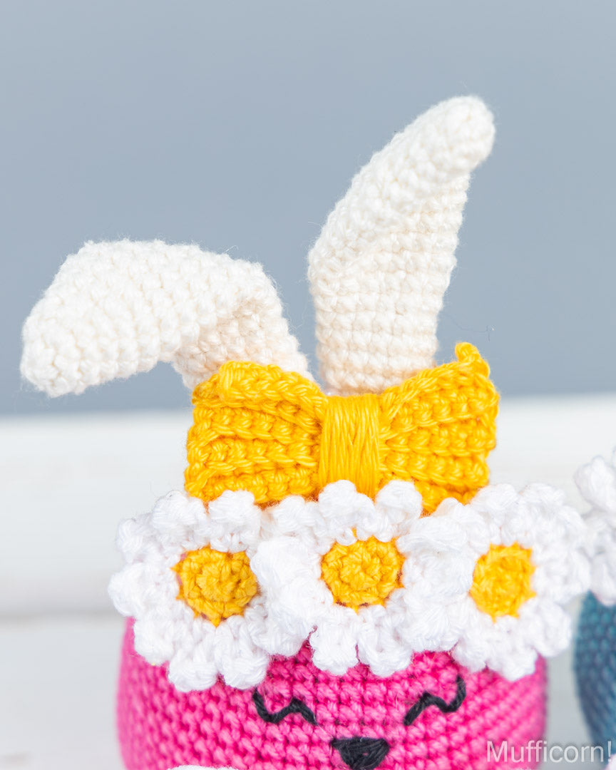 A close-up of a crocheted bunny character with white yarn ears, a yellow bow, and white daisy flowers around a pink face, shown against a soft gray background.